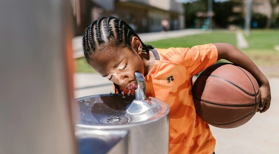 Little boy drinking water from the water fountain while holding a basketball