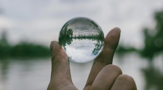 Fingers holding a transparent sphere reflecting the lake behind it